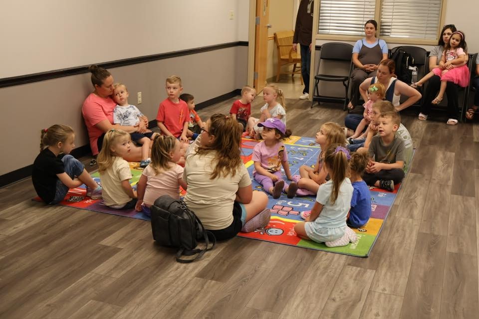 Children sitting in a circle on a colorful mat, with adults nearby in a room.