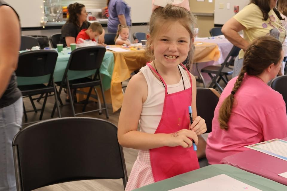 A young girl smiling, wearing a pink apron, holding markers in a room with others engaged in activities at tables.