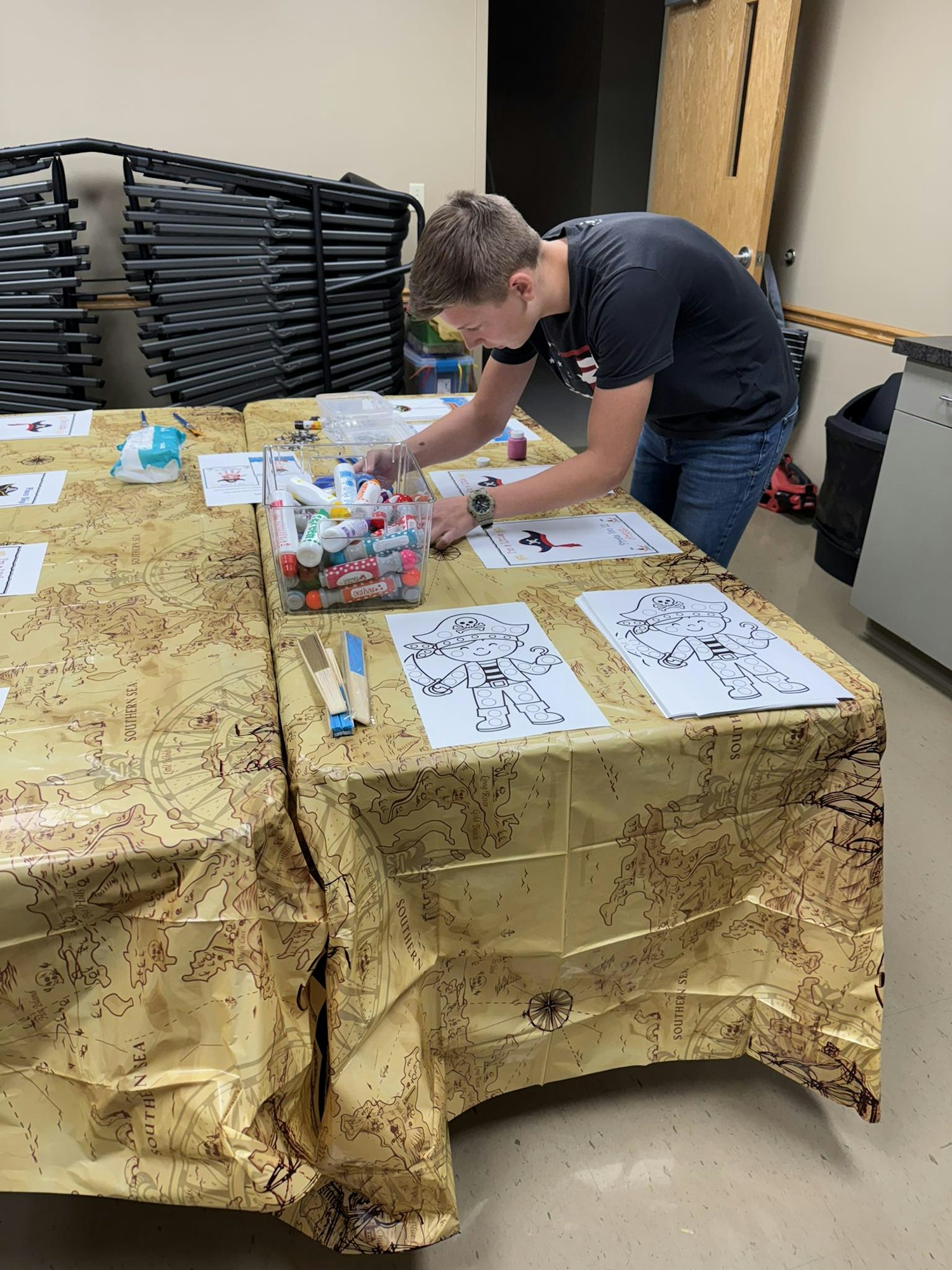 A person is setting up a craft station with coloring sheets and markers on a table with a map-themed tablecloth.