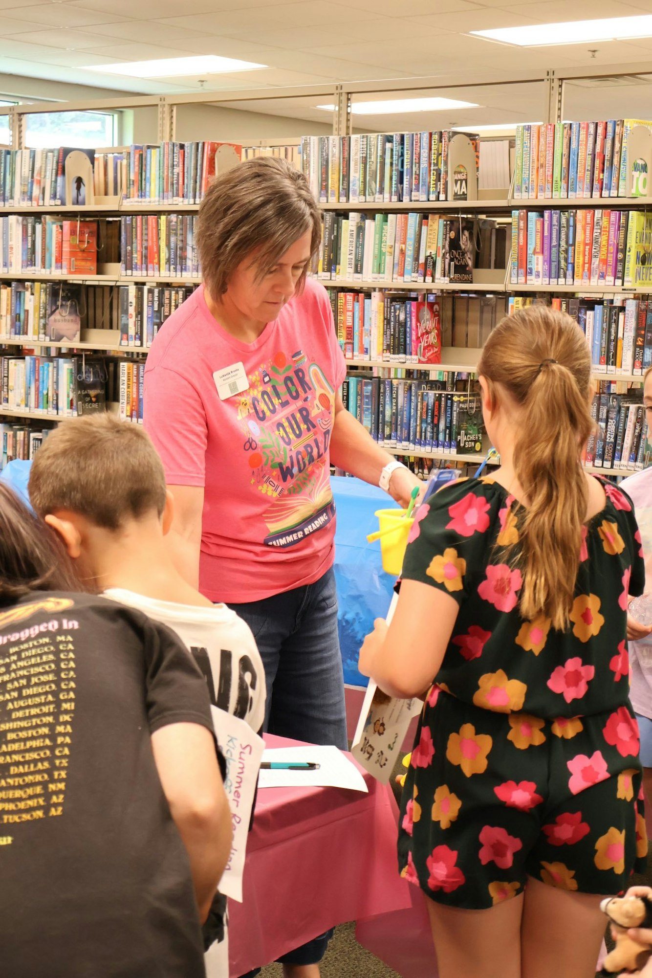 A woman at a library event interacts with children at a table. Books are displayed on shelves in the background.