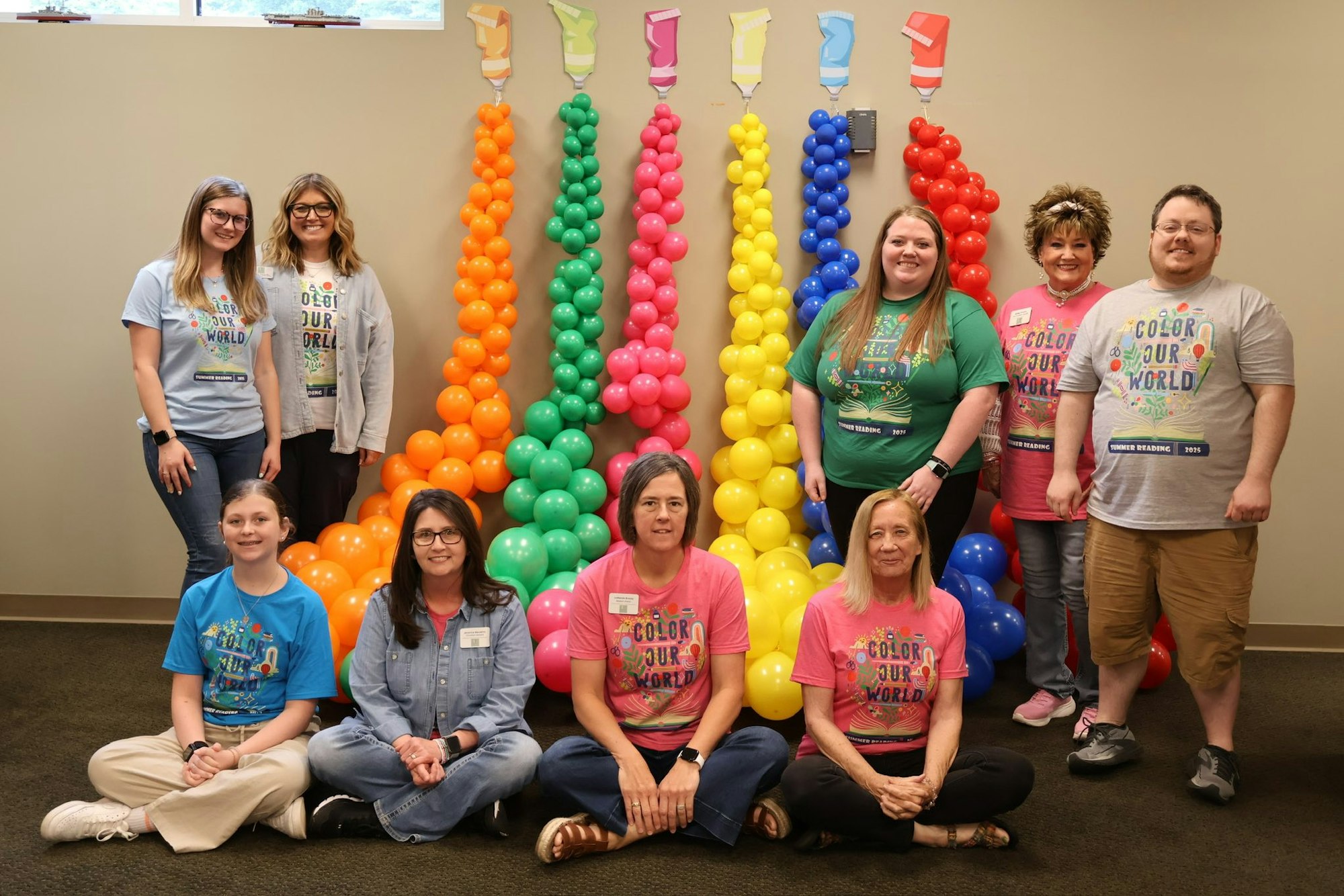 A group of people in colorful shirts poses in front of rainbow balloon columns.