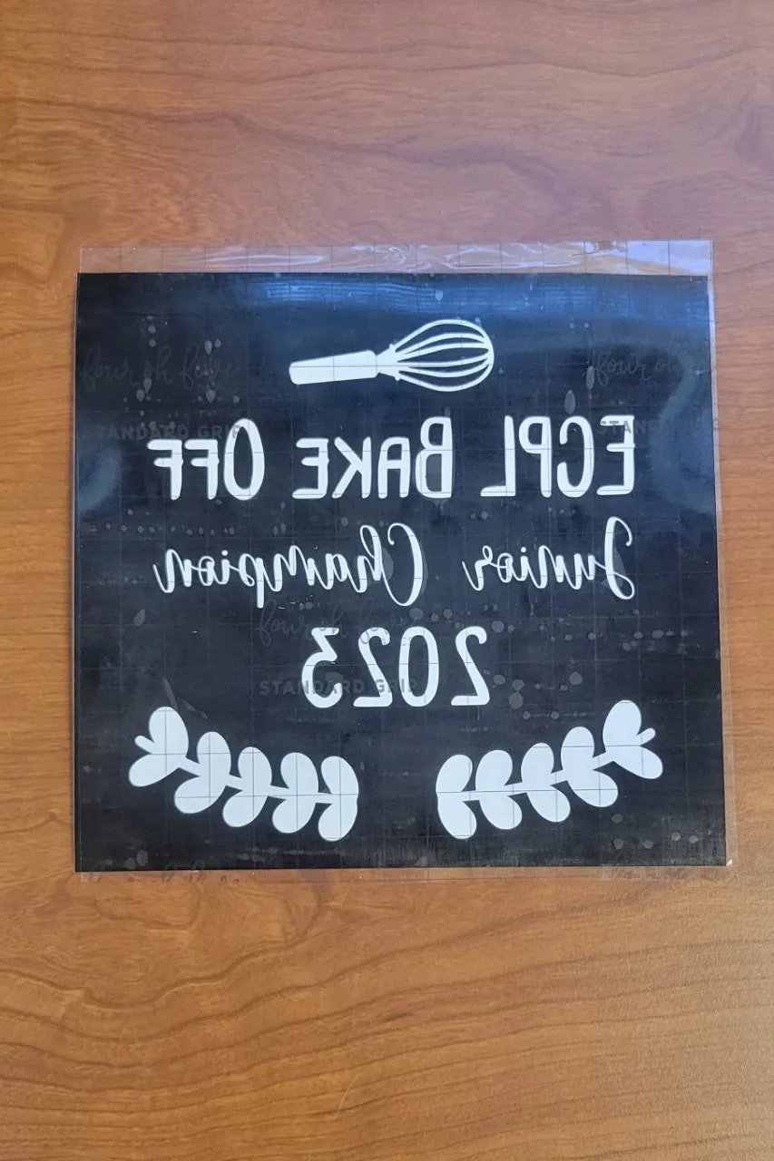A sign reads "Epic Bake Off Junior Champion 2023" with a whisk and leaves design, view is mirrored.