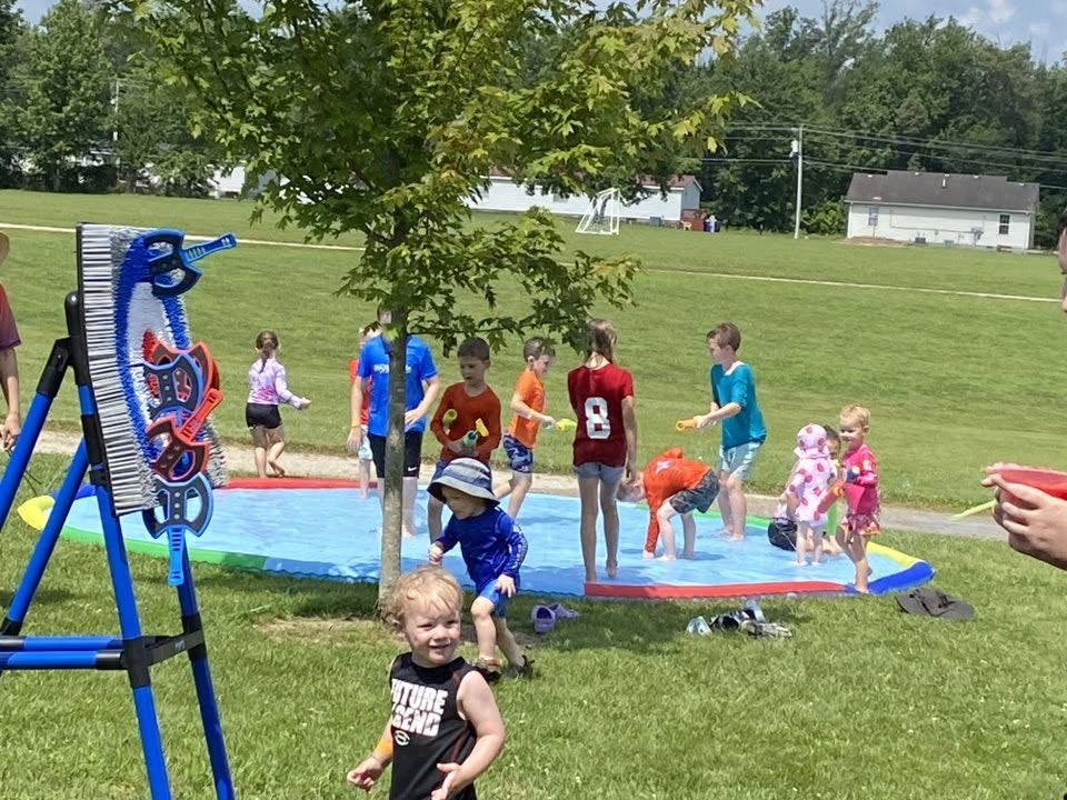 Kids playing on a splash pad in a park, with water toys and a tree nearby.