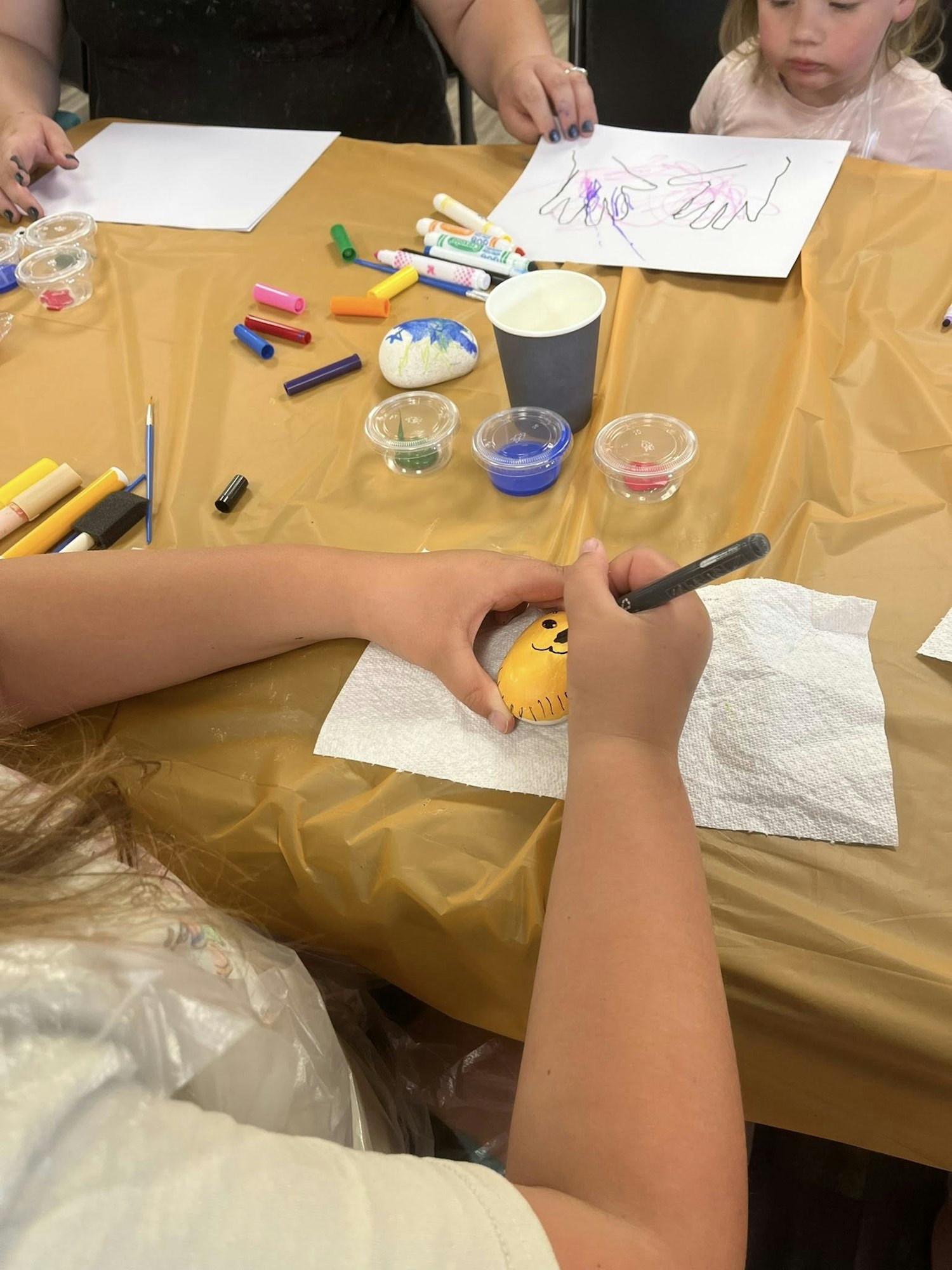 A child decorates a rock with markers at a craft table, surrounded by art supplies.
