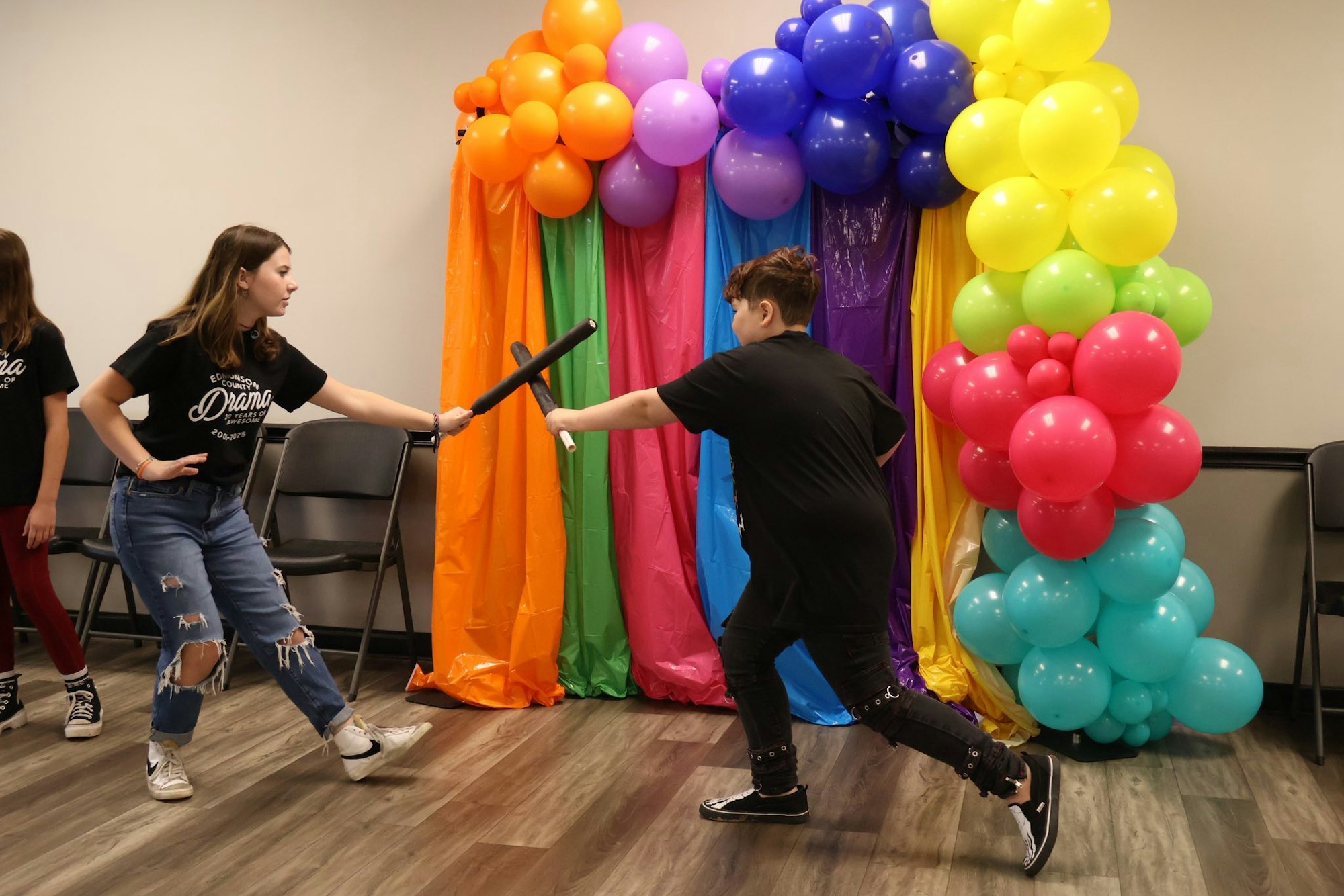 Two kids are play-fighting with foam swords in front of a colorful balloon and curtain backdrop.