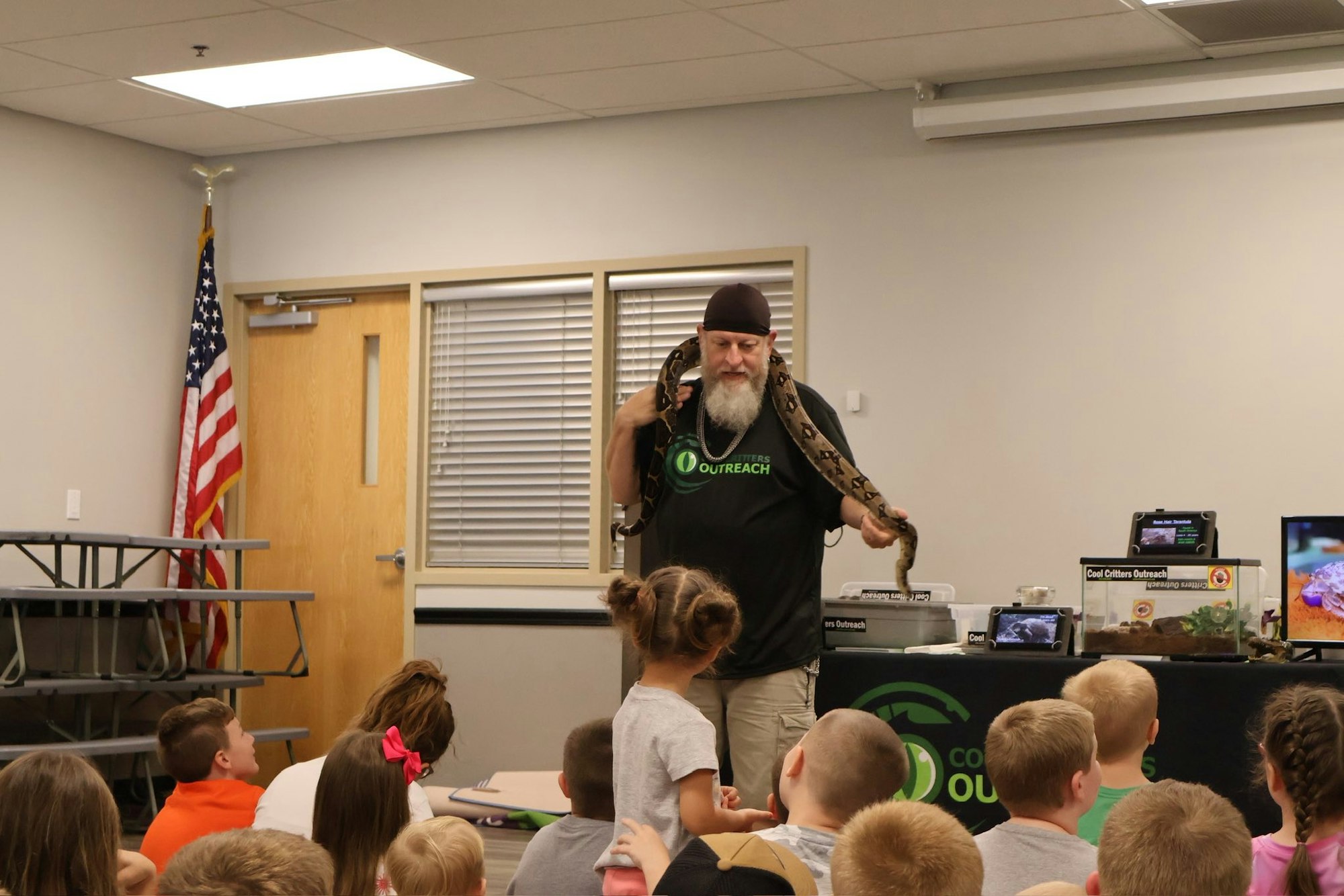 A man with a snake on his shoulders presents to children in a classroom setting.
