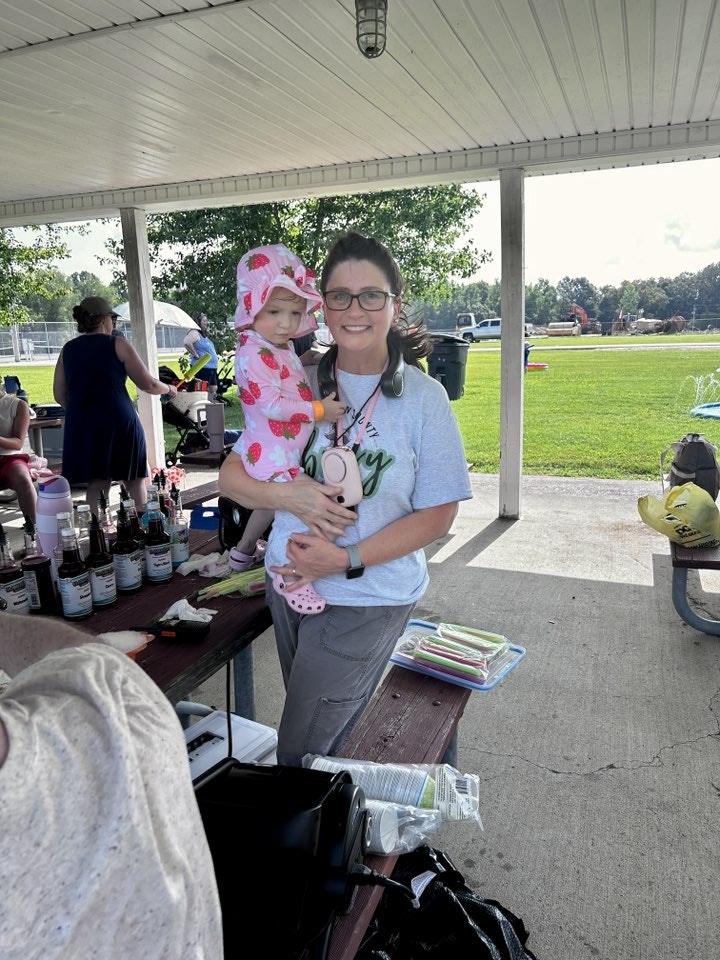 A woman holding a child in a strawberry-patterned outfit under a pavilion near a park, with various items on a table.