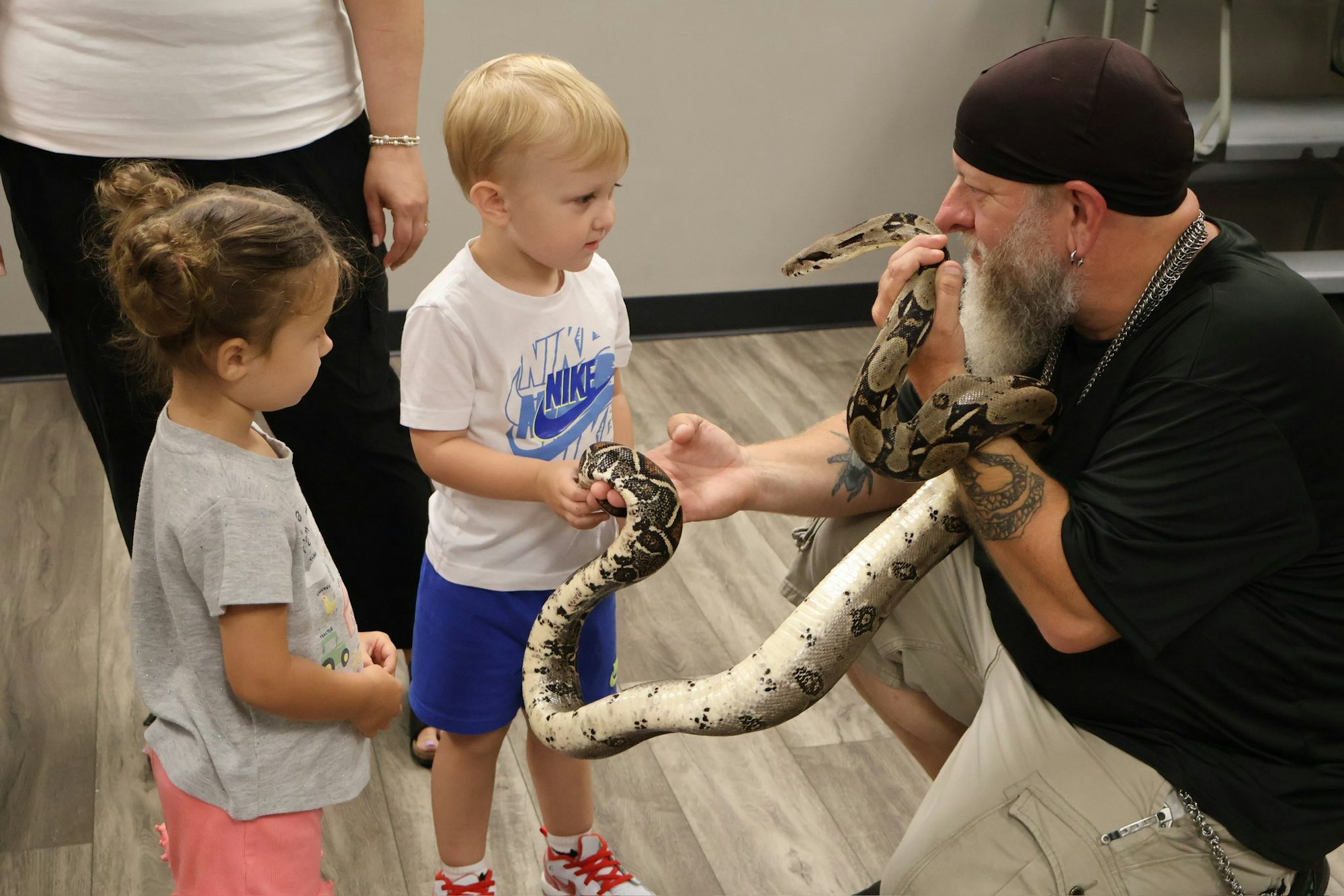 A man shows a large snake to two children, as they curiously observe and interact with it.