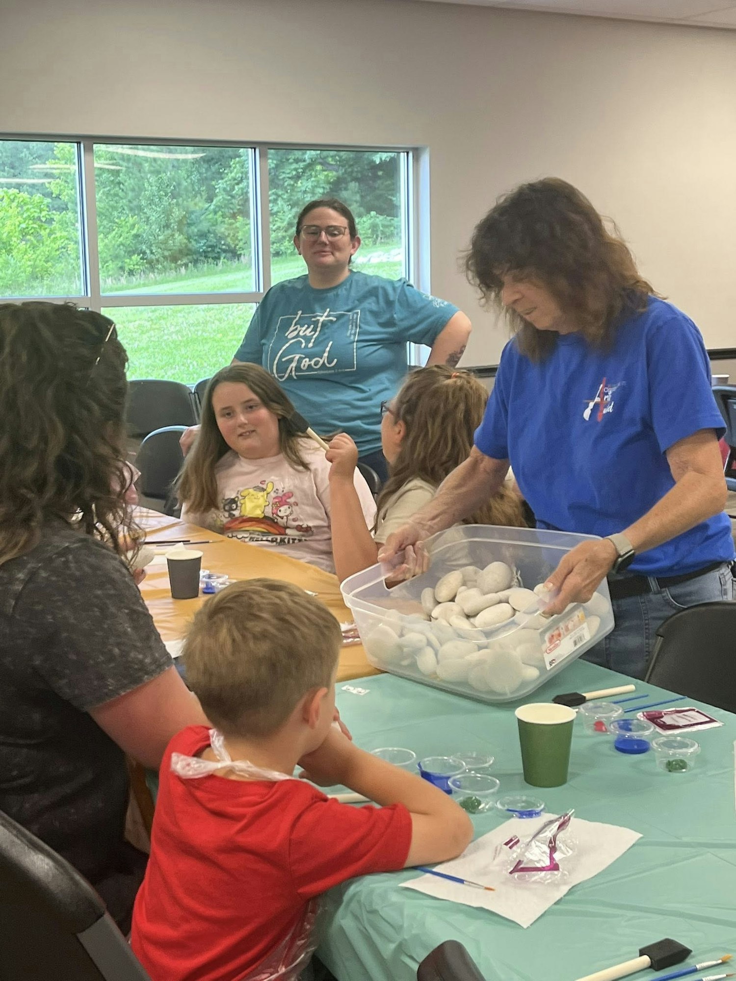 A group of adults and children engage in a craft activity with rocks and paint at a table indoors.
