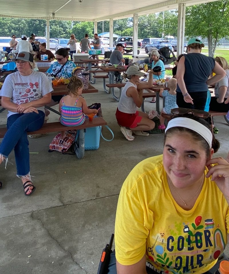 People sitting and standing under a pavilion, eating and socializing with various bags and coolers around.