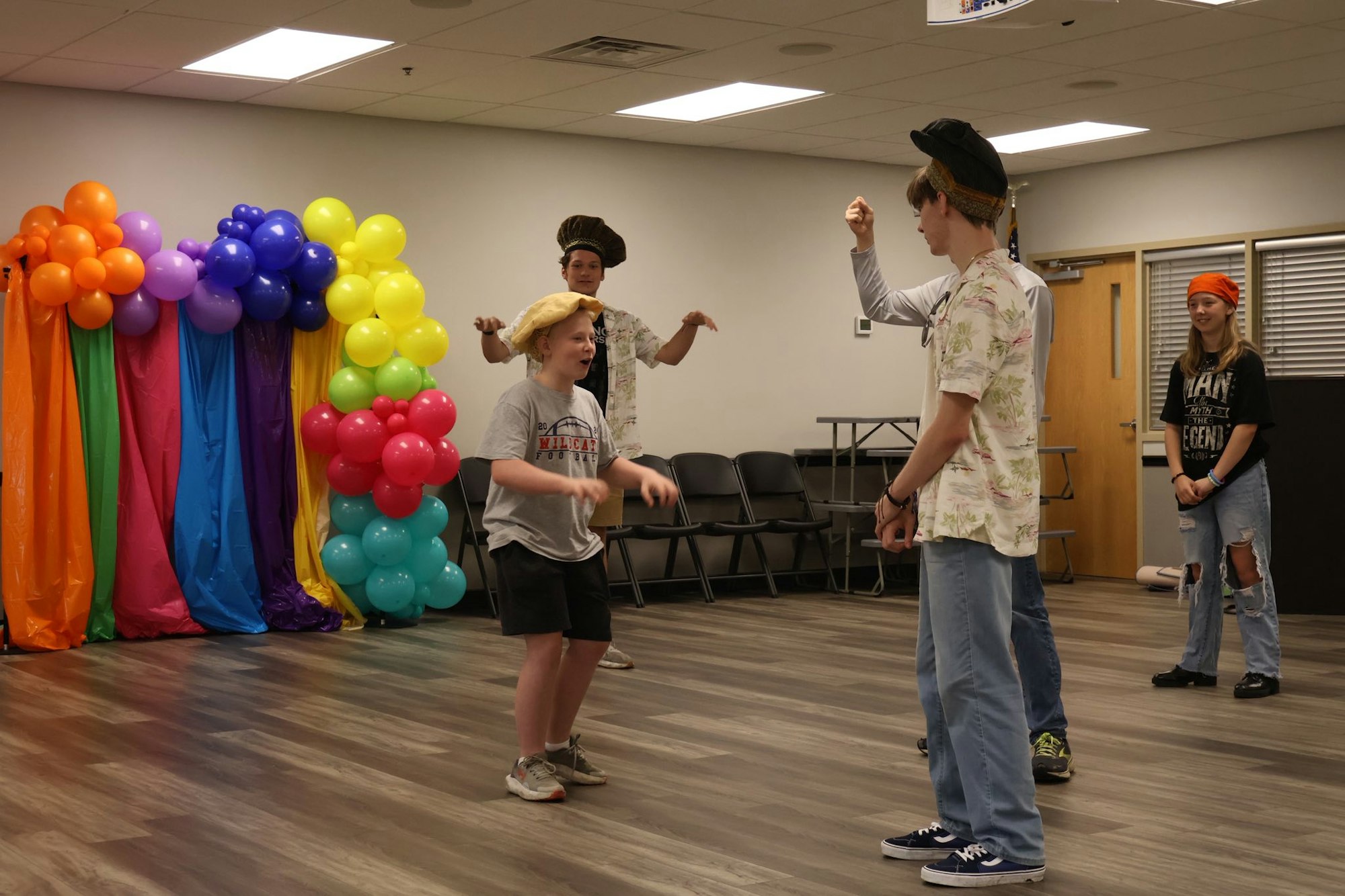 A group of people, some in hats, are engaging in an activity near a colorful balloon arch indoors.