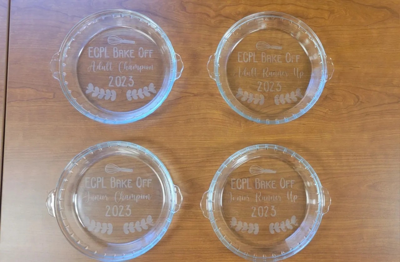 Four glass pie plates with "ECPL Bake Off" awards for Adult/Junior and Champion/Runner-Up 2023, on a wooden surface.