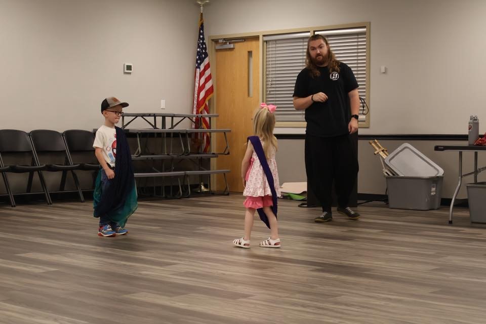 Two children in capes facing an adult in a room with folded chairs, an American flag, and storage bins.