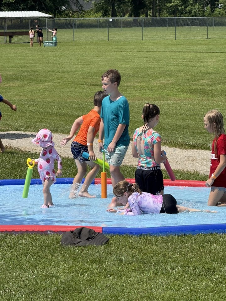 Kids playing in a splash pool with water toys on a sunny day in a grassy field.