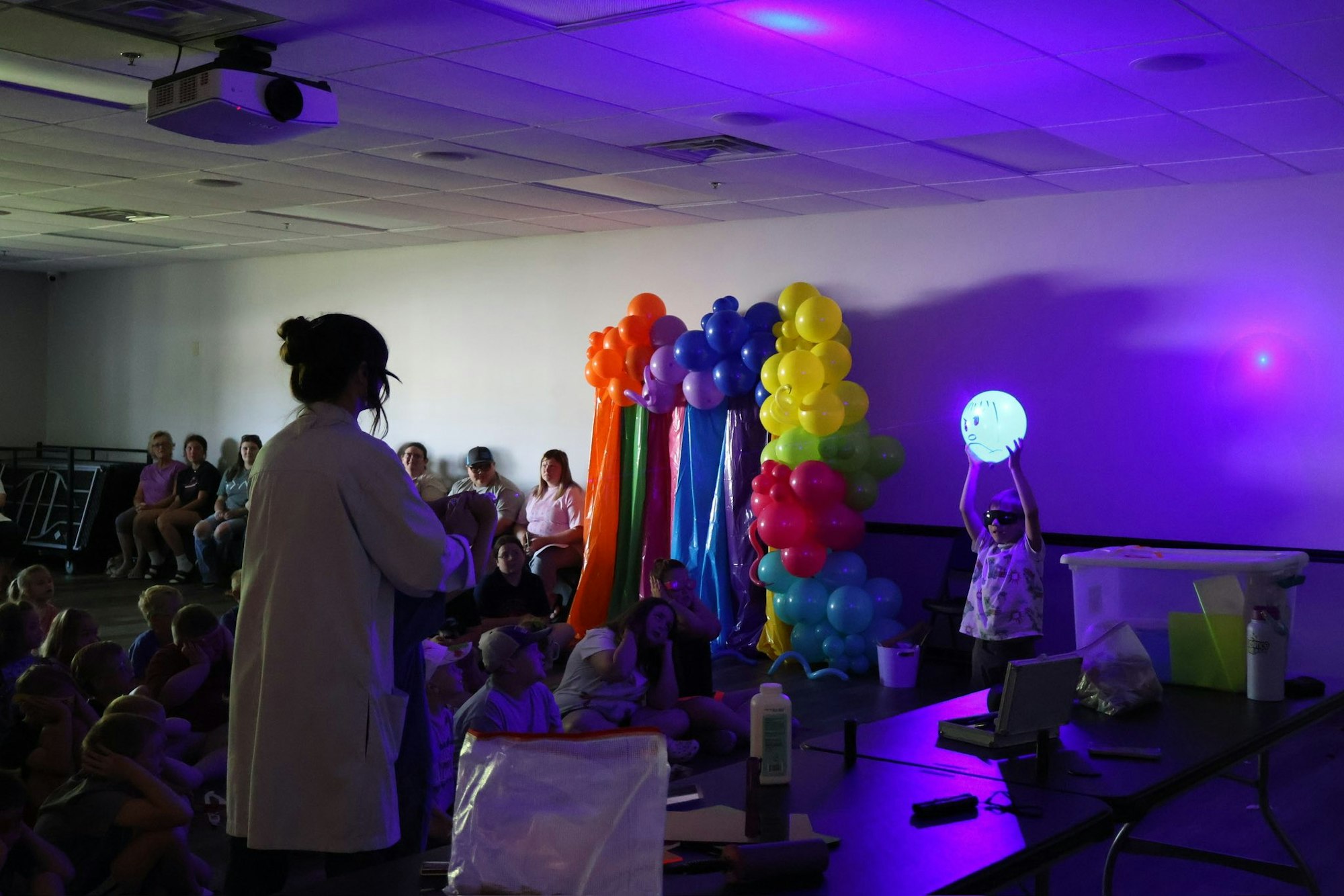 A person in front of an audience with a balloon arch, lit by purple lights. A child holds a glowing ball, wearing sunglasses.
