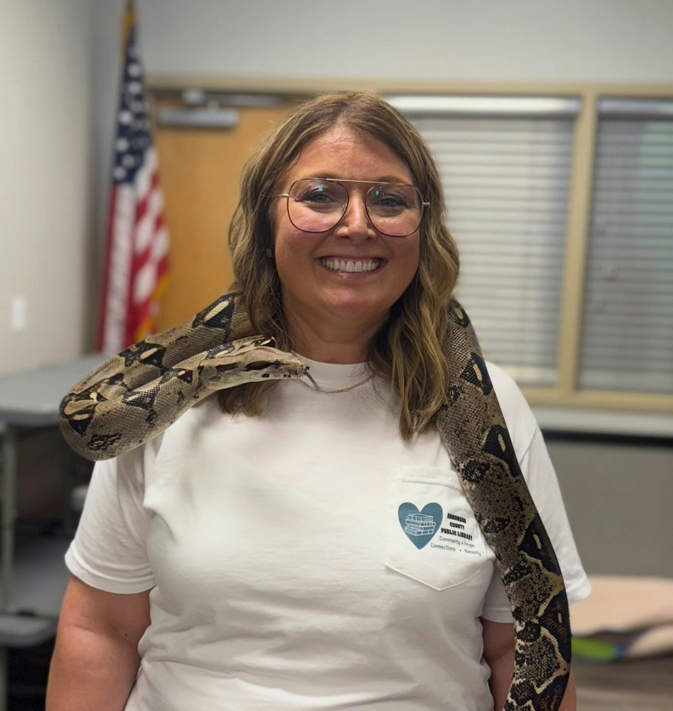 A person smiling with a snake around their shoulders, standing indoors with an American flag in the background.
