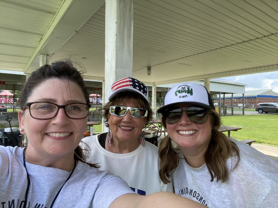 Three smiling people wearing caps and sunglasses under a pavilion.