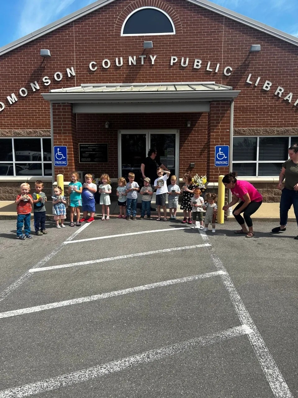 A group of children and adults stand outside Edmonson County Public Library, near a ramp and accessible parking signs.