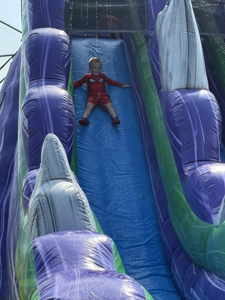 A child in a red outfit slides down a colorful inflatable slide outdoors.