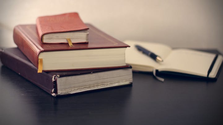 Stacked books with bookmarks on a desk, next to an open notebook and a pen.