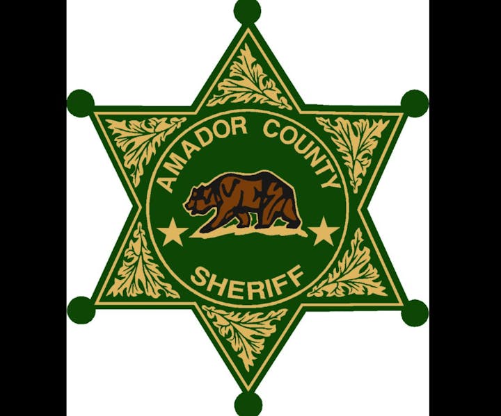 A green and gold badge emblem with "Amador County Sheriff" and a bear illustration.