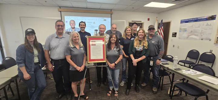 A group of people poses with a framed document, likely celebrating an event or recognition, in a meeting room.