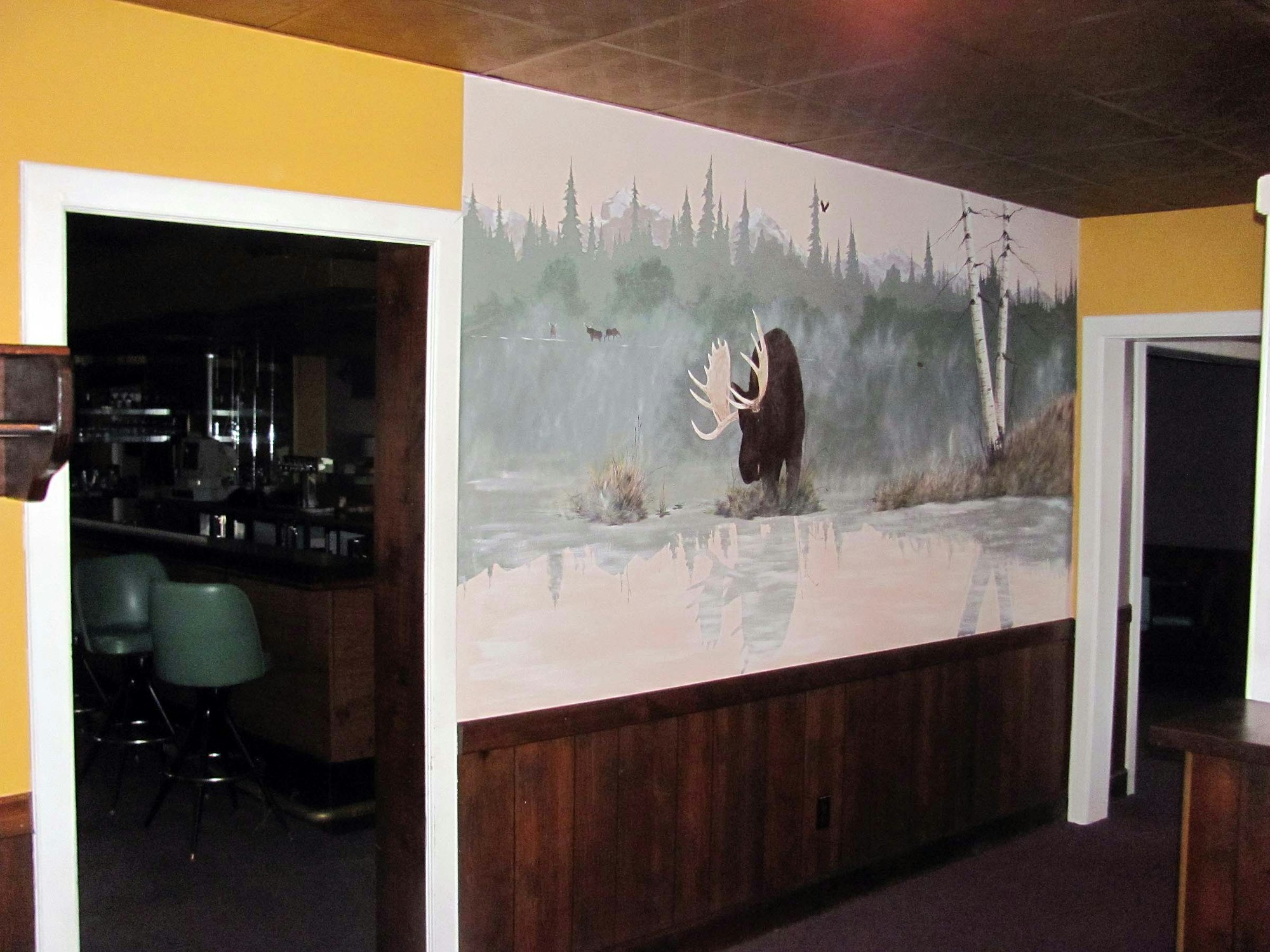 A mural of a moose by a lake in a forest setting; interior doorway and vintage bar seating visible.
