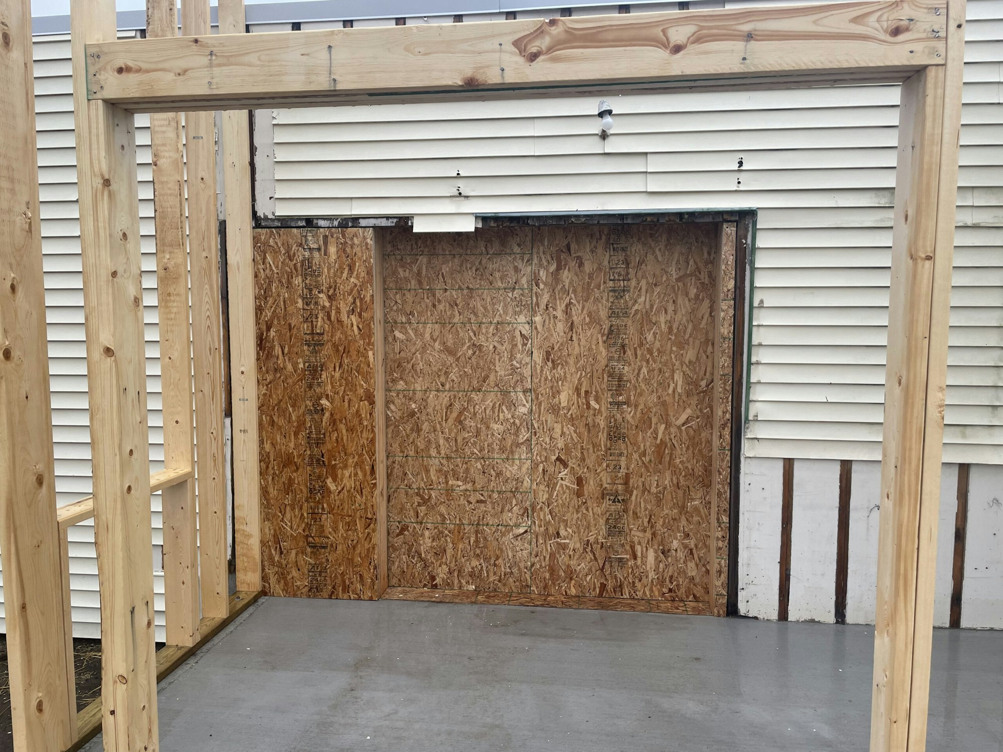 Wooden framing in front of a building with siding, a boarded-up doorway, and concrete flooring.