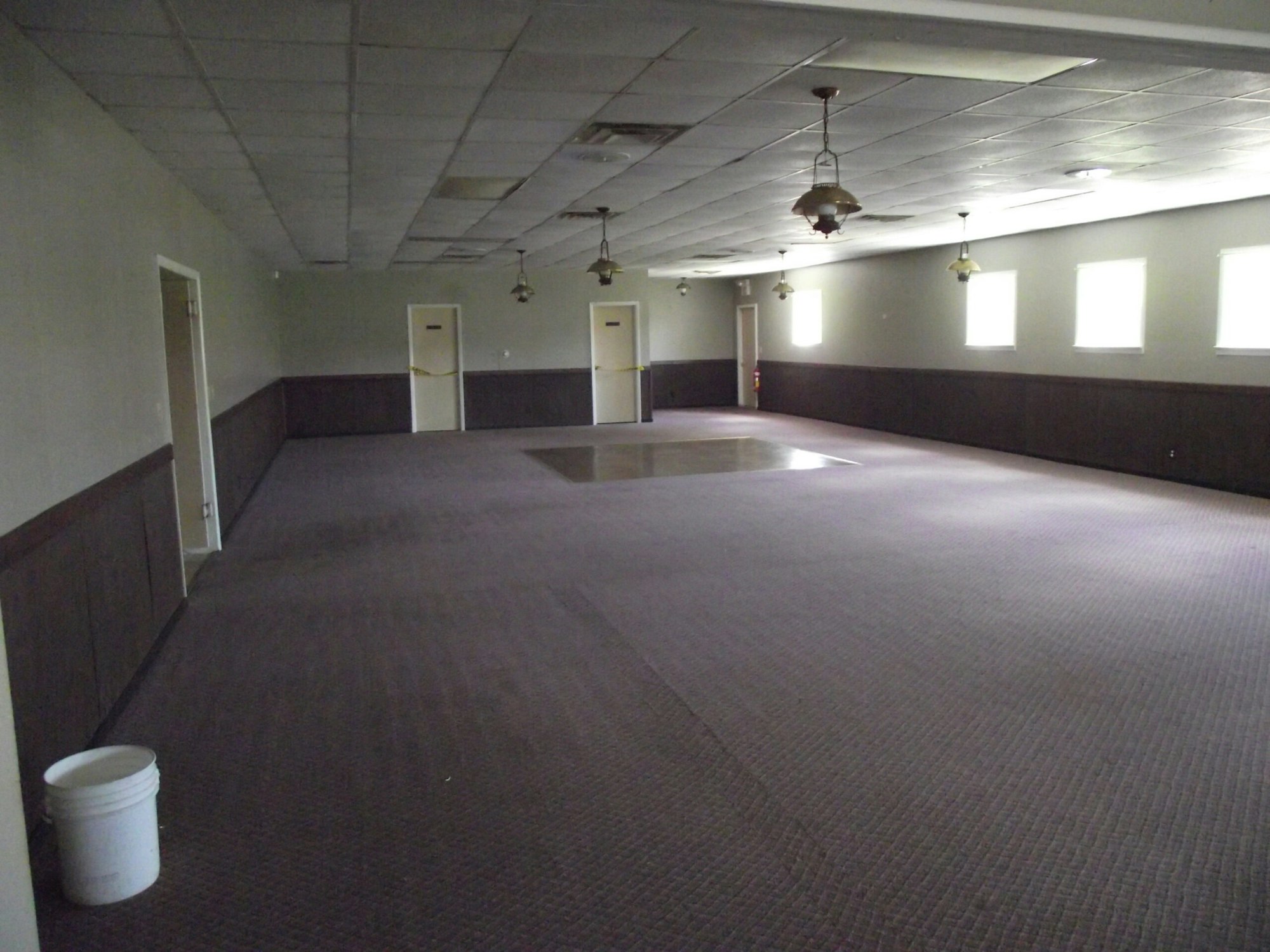 An empty room with carpet, ceiling lights, a white bucket in the foreground, and several doors and windows in the background.