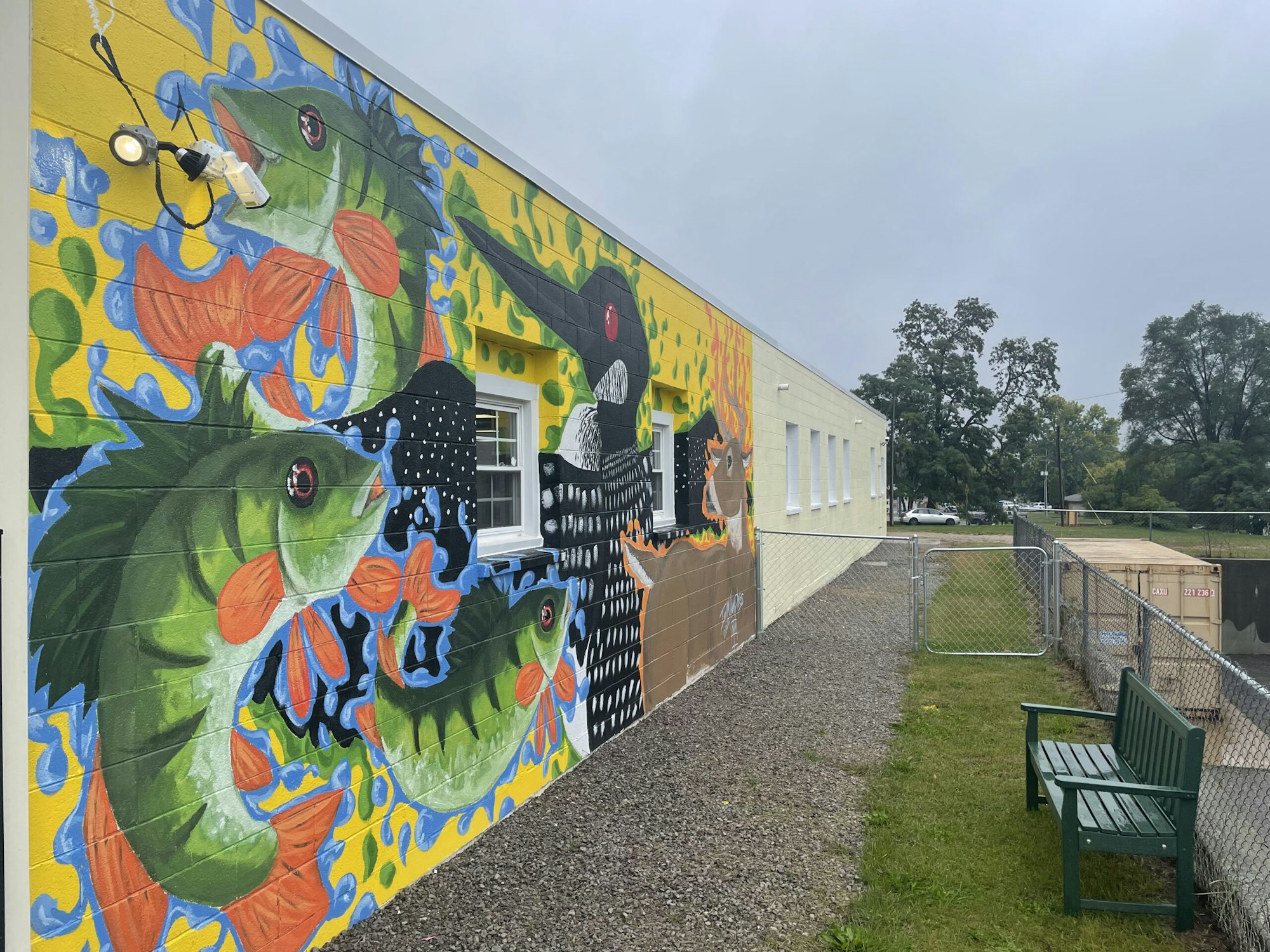 Colorful mural with fish and abstract designs on the side of a building; a bench and fenced area nearby.