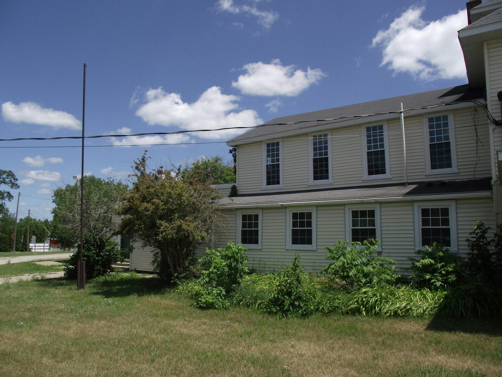A two-story beige house with multiple windows, surrounded by greenery and trees, under a blue sky with scattered clouds.