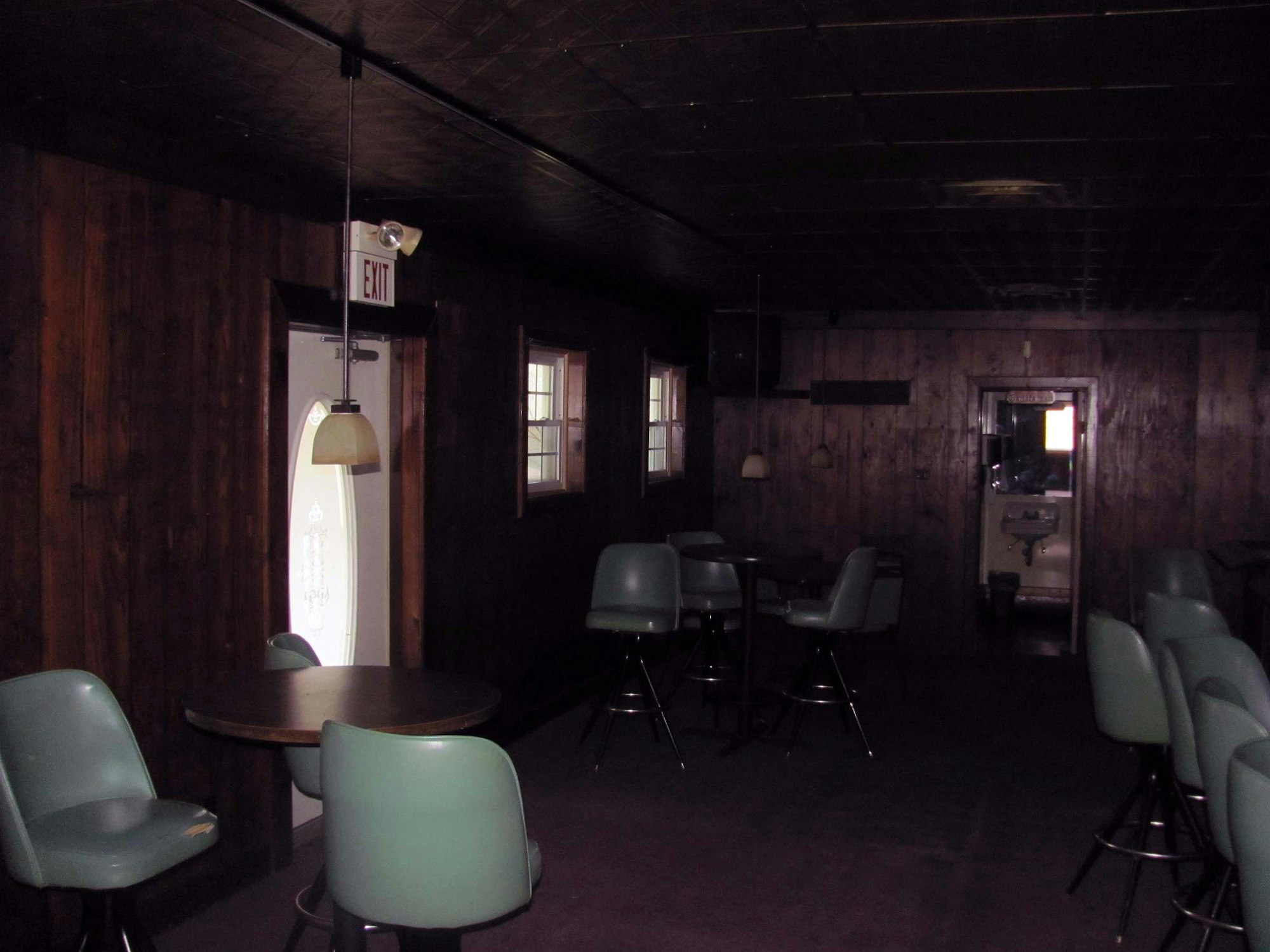 Dimly lit room with tables, chairs, and wooden walls. There's an exit sign above a door and some pendant lights.