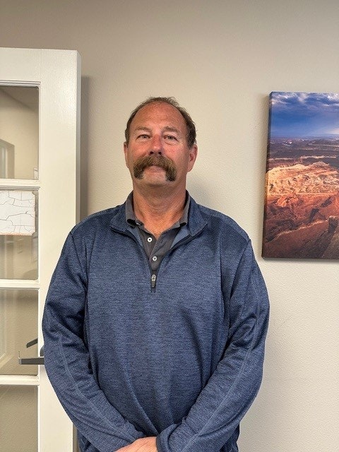 A person with a mustache wearing a blue sweater stands indoors, with a landscape painting on the wall behind them.