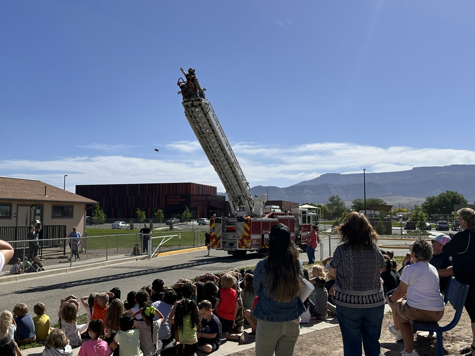 People watch a firefighter on a raised ladder next to a parked fire truck, with a clear blue sky and mountains in the background.
