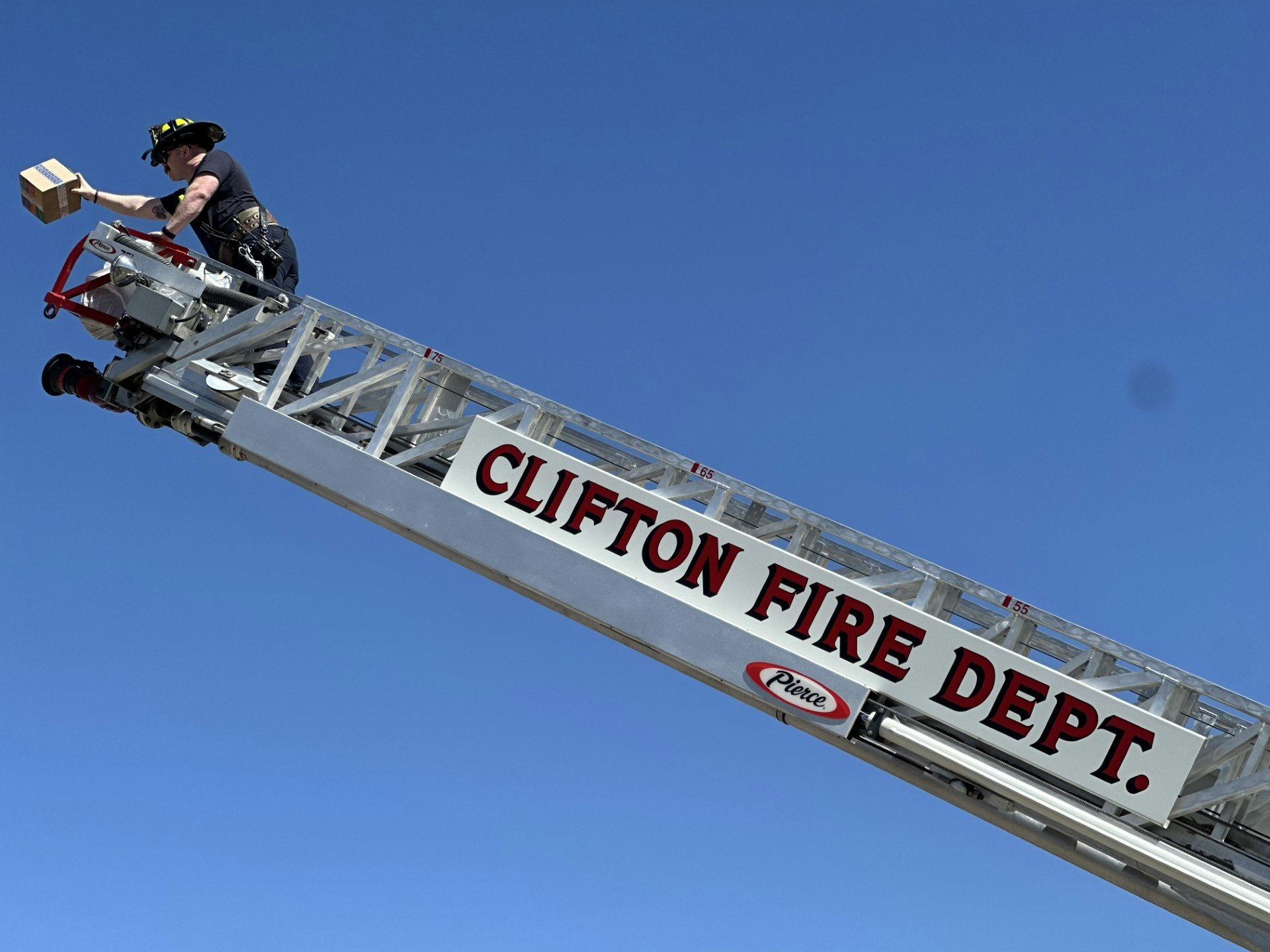 A firefighter on a ladder from "Clifton Fire Dept." holding a box, against a clear blue sky.