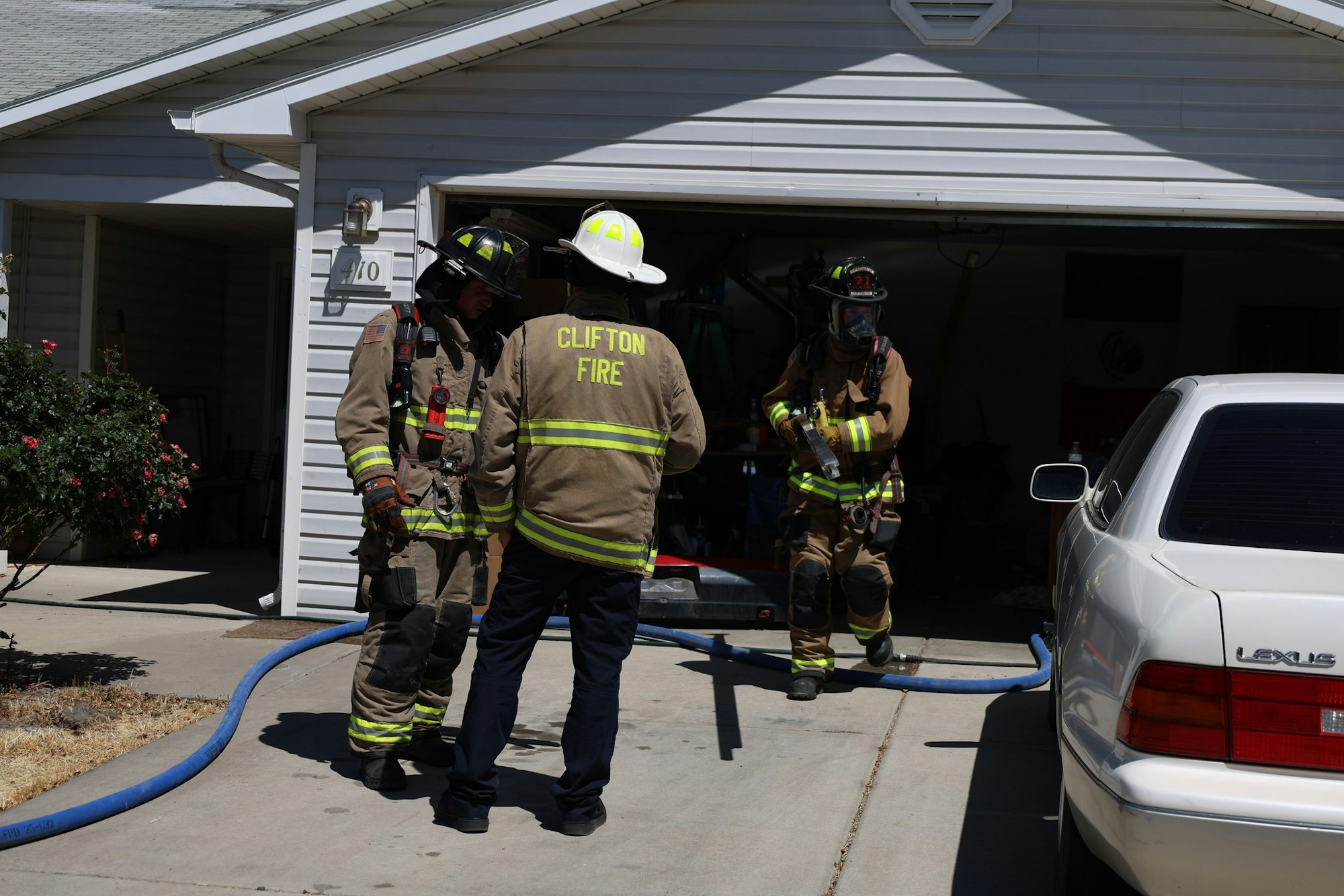 Firefighters in action, discussing a situation in front of a garage, with a fire hose and a white car nearby.