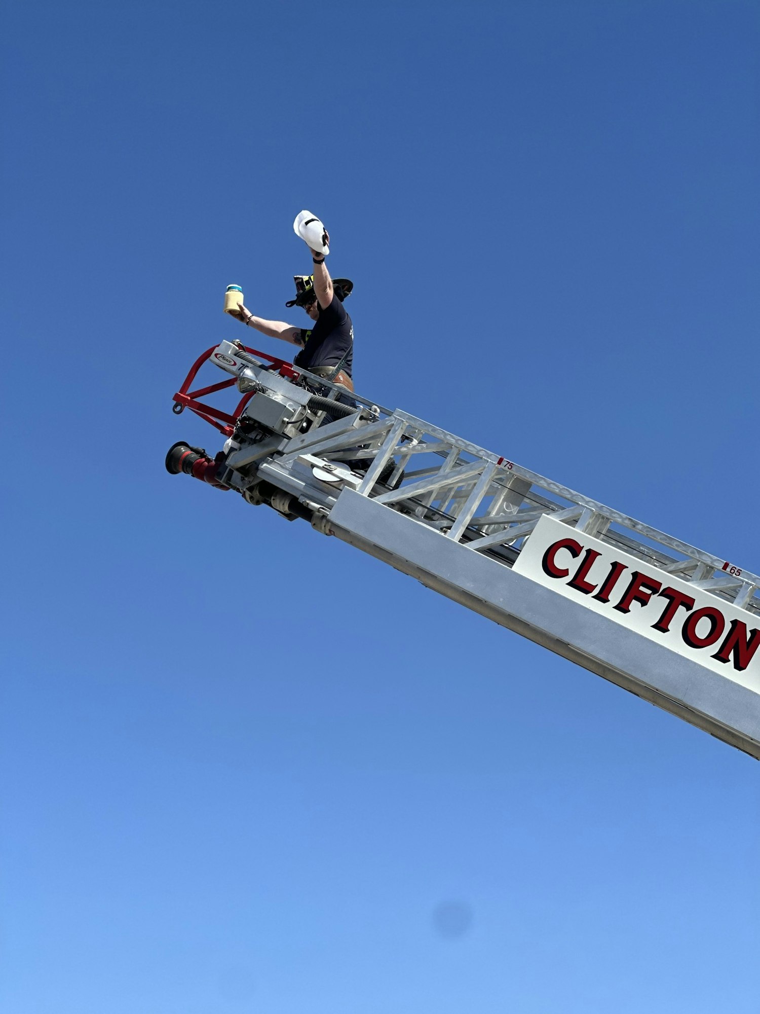 Person on a fire truck ladder holding a jar and a hat under a clear blue sky.