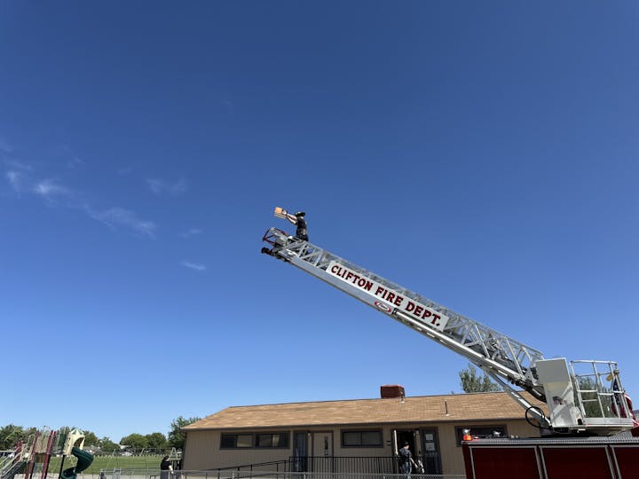 Firefighter on a ladder truck holds a book high under a clear blue sky, labeled "CLIFTON FIRE DEPT."