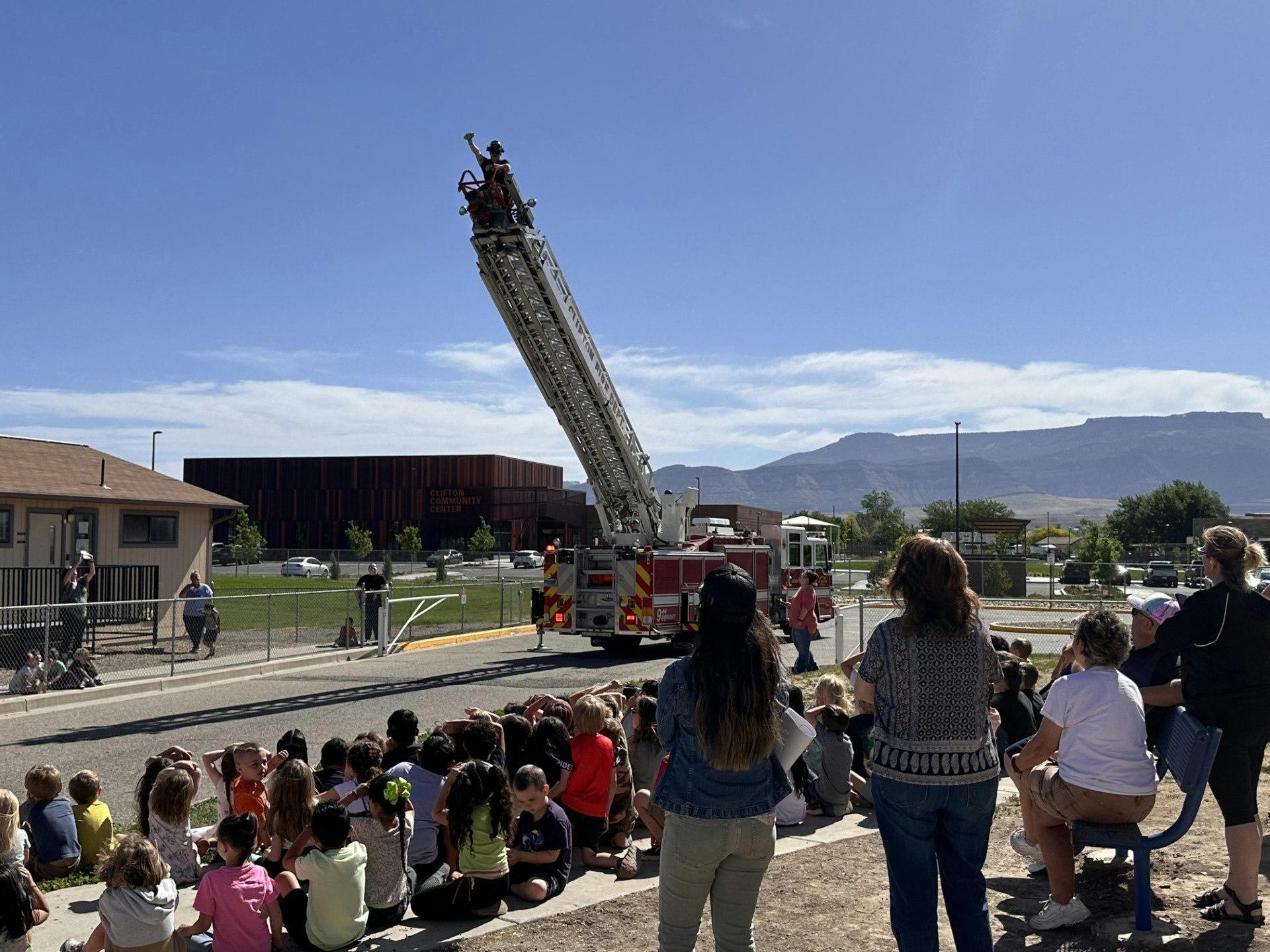 A fire truck with an extended ladder entertains a seated group outdoors.