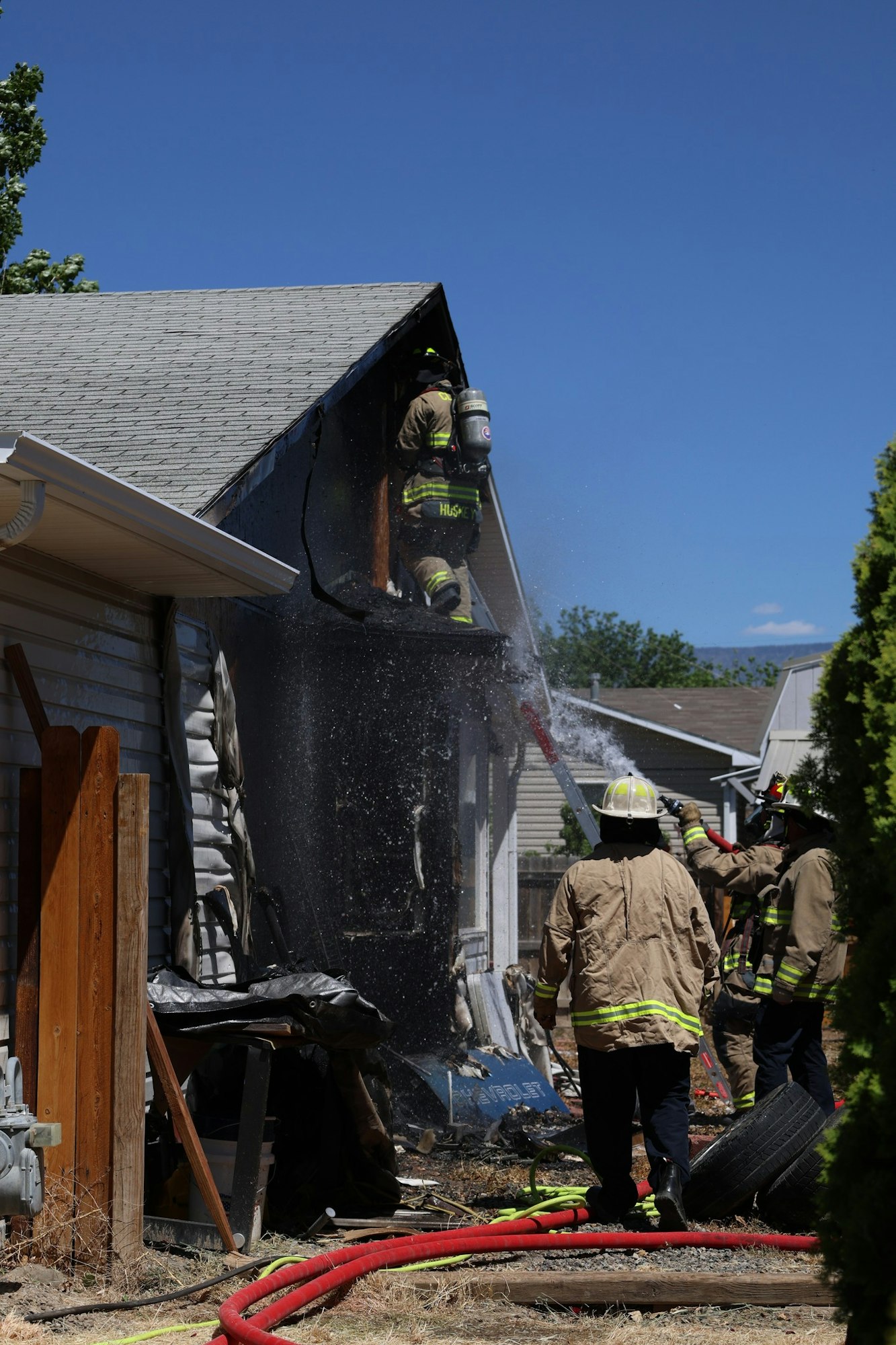 Firefighters are battling a house fire, using hoses and equipment to extinguish flames near a damaged building.