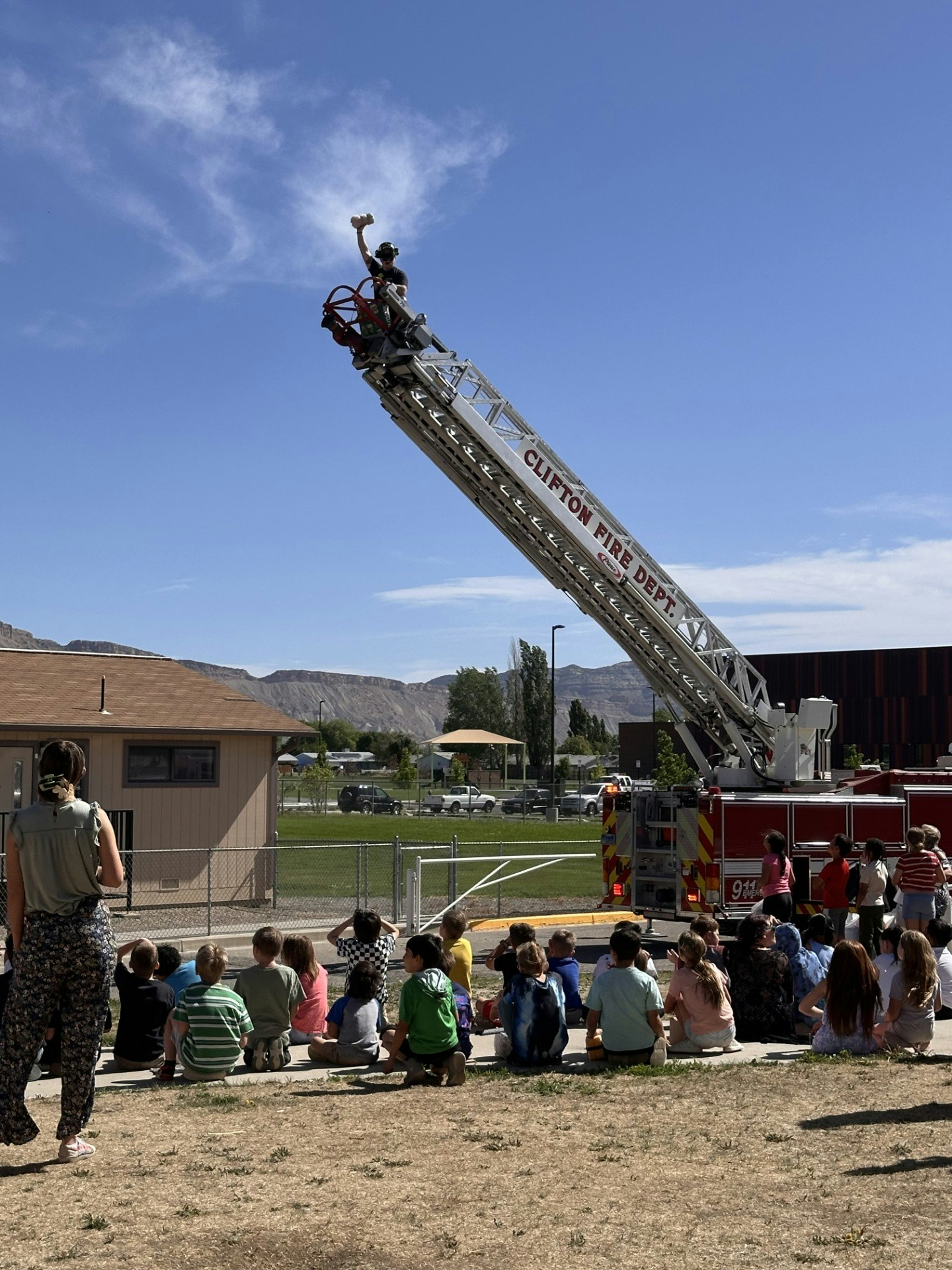 Firefighter on a ladder truck, holding an object, with an audience of children and adults seated on the ground watching.