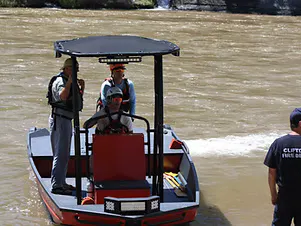 Three people on a small boat with a canopy in a river, one person standing nearby onshore.