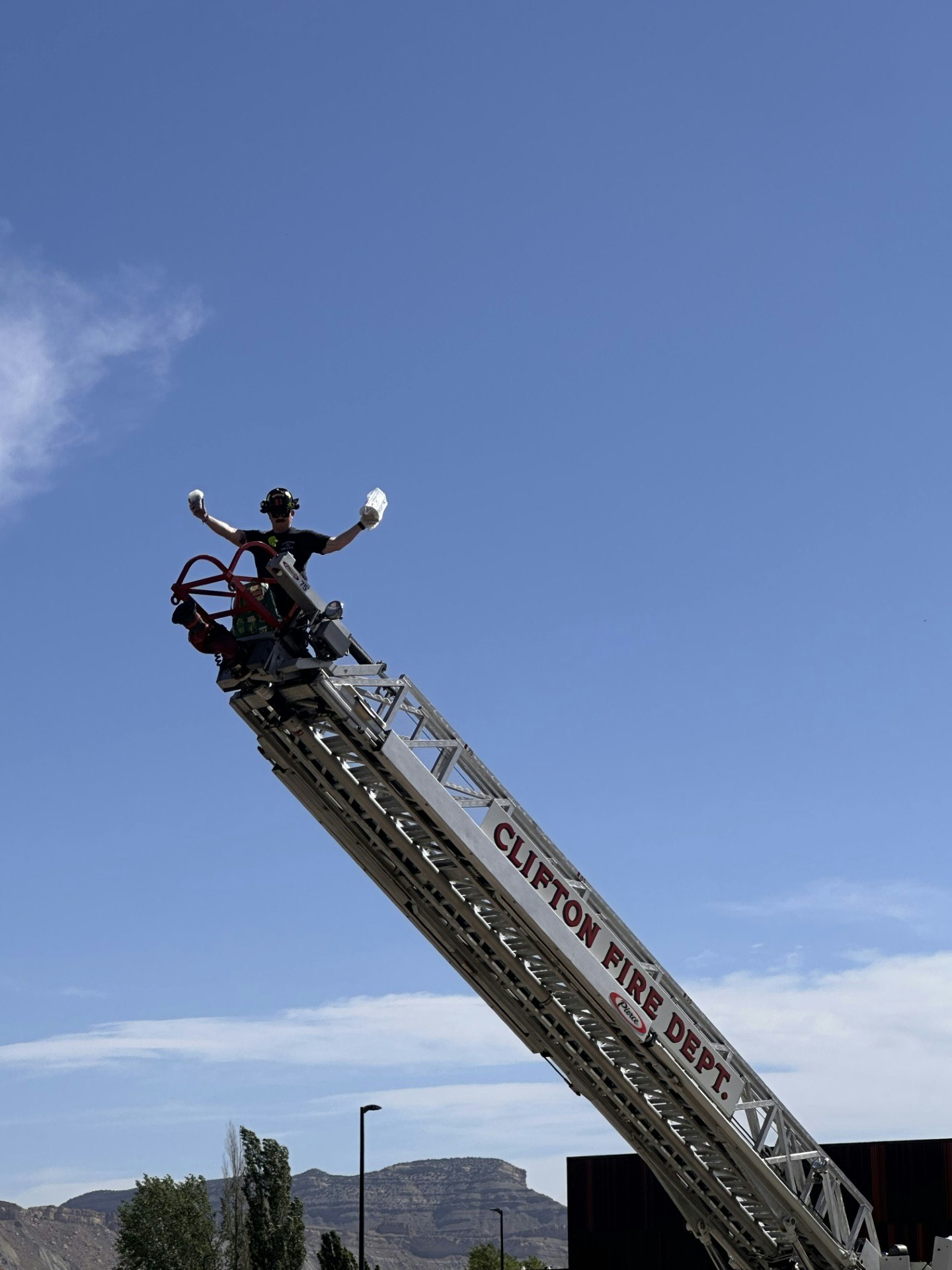 Firefighter on a raised ladder, holding items aloft, with clear blue sky background, "Clifton Fire Dept." visible on ladder.