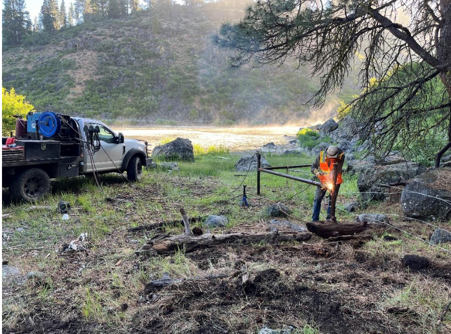 A person in safety gear is welding near a river, with a utility truck and equipment nearby, surrounded by nature.