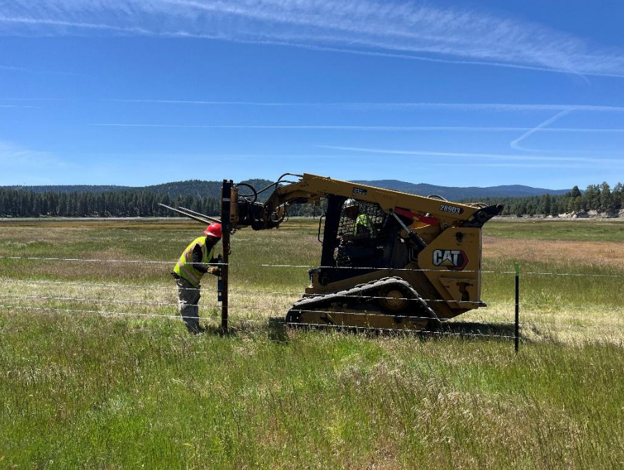 A person in a safety vest and helmet works with machinery in a grassy field setting up fencing.