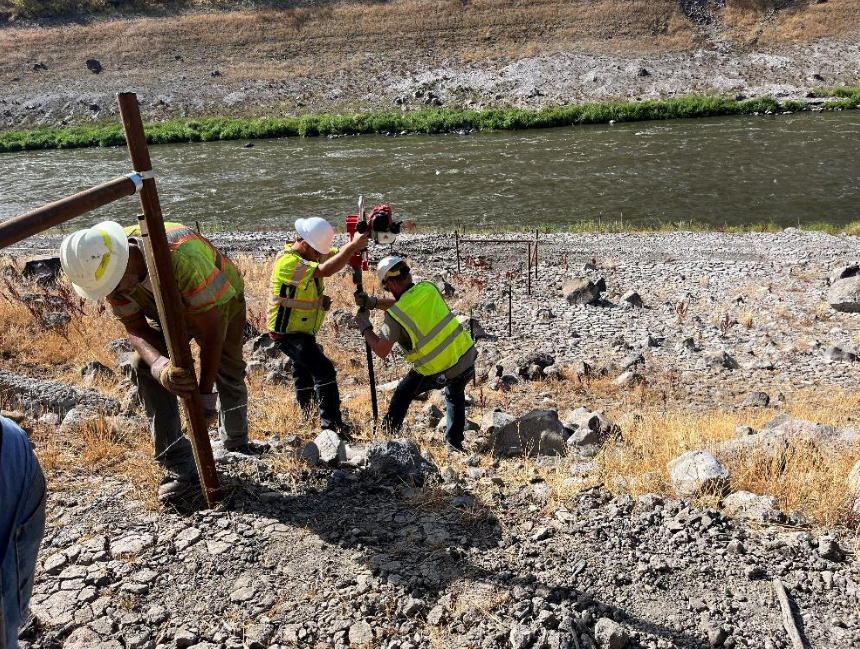 Construction workers in safety gear install a fence post near a riverbank.