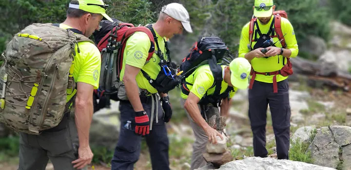 A group of hikers in bright yellow shirts and backpacks examines a rock in a natural setting. They appear to be in a forested area.