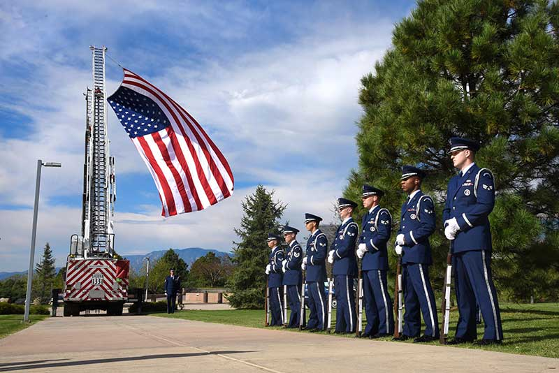 Uniformed military personnel stand in formation as a large American flag is displayed on a fire truck ladder.