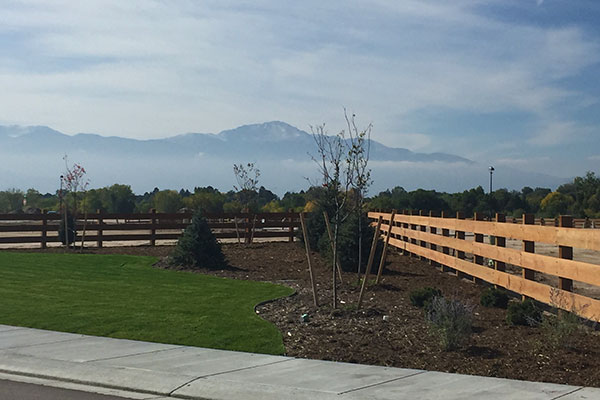 A manicured landscape with a wooden fence, small trees, and mountains in the background under a partly cloudy sky.