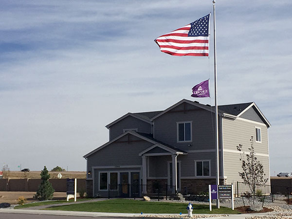 A two-story house with a U.S. flag and a purple flag on flagpoles outside, under a clear sky.